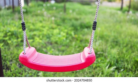 An empty lonely red swing hangs in a garden full of flowering trees and green grass in a warm springtime.