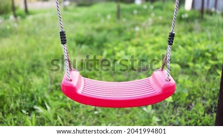 An empty lonely red swing hangs in a garden full of flowering trees and green grass in a warm springtime.
