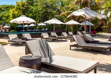 Empty Lounge Chairs With Umbrellas On White Sand Beach In Hot Summer Day In Luxury Tropical Hotel Or Resort. Travel, Tourism, Holiday, Vacation Concept. Palm Trees, Greenery, Swimming Pool Background.