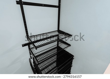 An empty metal storage cage with multiple stacked wire shelves stands indoors against a plain white background. The black steel frame and rigid grid panels create a structured industrial look, with soft even lighting and no people or products inside