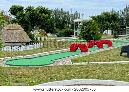 Empty miniature golf course on a cloudy summer day, featuring green tracks, bright red bridge obstacles, small decorative thatched house, trees, and white fences. Perfect for summer recreation, family vacation, and outdoor activities concepts.