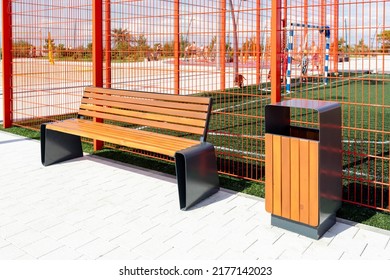 An Empty Modern Bench And A Trash Can Near A Sports Football Field With A Red Metal Fence On A Sunny Summer Day. Renovation Of Territories, Modern Design And Style Of City Landscape