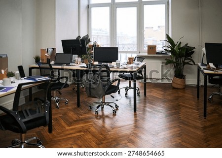 Empty modern office showing multiple workstations with computer monitors, chairs, scattered office supplies, potted plant in corner, clean hardwood floor, large windows providing natural light