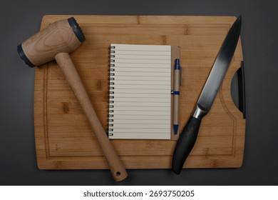 Empty notebook on bamboo cutting board with kitchen tools.

