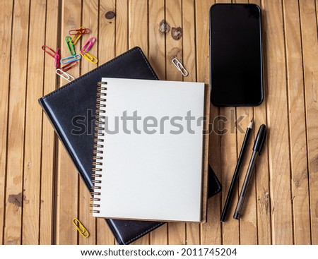Empty Opened Journal Beside Mobile With Pens On Top Of Wooden Desk. Blank Spiral Notebook Pad Alongside A Phone With Ballpens Placed On A Table.