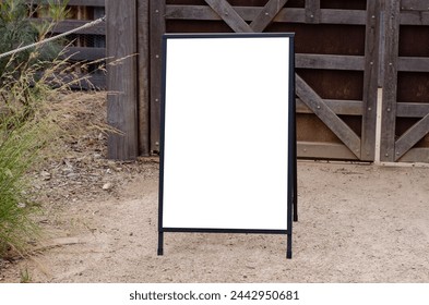 Empty outdoor advertising stand against a rustic wooden background. Blank white mockup template of a freestanding sandwich board at a farm gate. Copy space for your design.