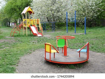 Empty Outdoor Playground For Children With A Slide, Carousel, Pull Up, Chin Up Bar In The Park With Blooming Trees In The Background.