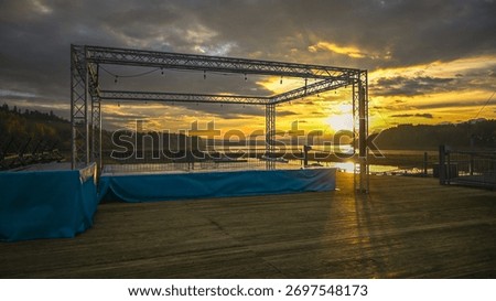 Empty outdoor stage with truss structure overlooking a lake at sunset.
