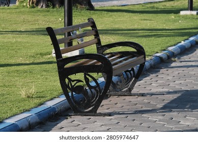 Empty Park Bench Along Paved Pathway Next To Green Lawn In Sunny Urban Park