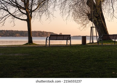 Empty park bench in front of an idyllic lake. The sun is going down and the sky is glowing in orange color. Tranquil scenery without people. Landscape with a resting place in a lakeside area.