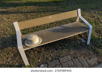 Empty park bench with straw hat resting on seat, bathed in warm sunlight evoking peaceful and relaxing mood outdoors in nature on quiet afternoon with shadows stretching across grass