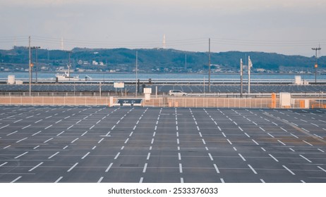 Empty parking space with lined asphalt, overlooking a calm waterfront and distant hazy hills. A ship sails near the coast under a dim sky. - Powered by Shutterstock