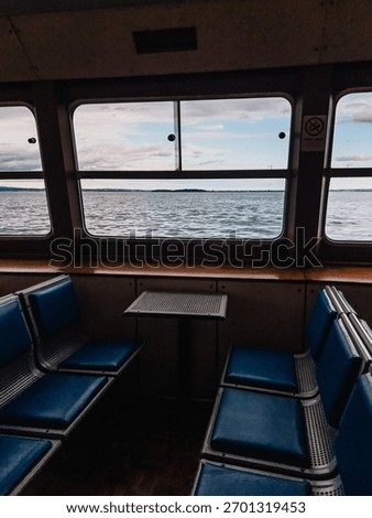 Empty passenger seats and small table inside ferry boat navigating on Lake with view on water and cloudy sky through large windows. Vacant ferry seats, small table, large windows revealing Lake waters