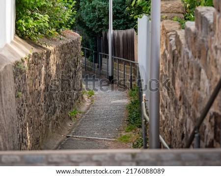 Empty path with big old stone walls and stairs downhill. Asphalt way without any people. Traditional exterior in Europe. Lush foliage in the summer season. Narrow footpath in a small mountain town.