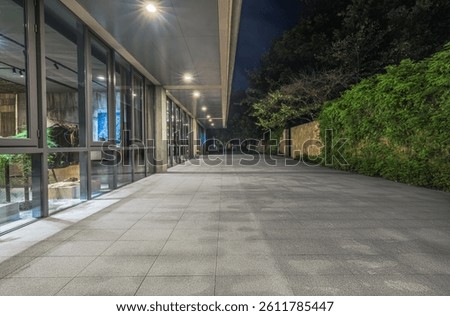 Empty paved square walkway alongside modern glass facade office building exterior illuminated at night with green plants.