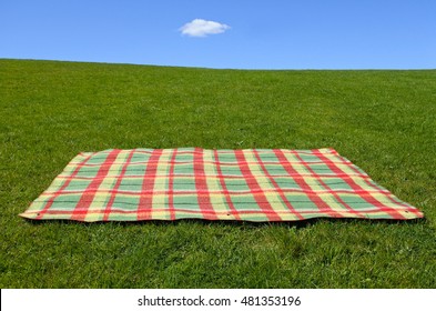 An Empty Picnic Blanket On Green Grass Under Blue Sky With One Cloud. Family Holiday Concept. No People. Copy Space