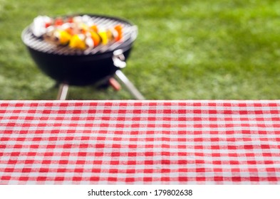 Empty picnic table background covered in a fresh country red and white checked cloth for your product placement or advertising with a barbecue on a green lawn behind