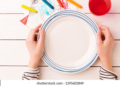 Empty Plate In Child's Hands On White Planked Wooden Background With Colorful Decorations. Table Setting For Kids - Captured From Above (top View, Flat Lay). Layout With Free Text (copy) Space.