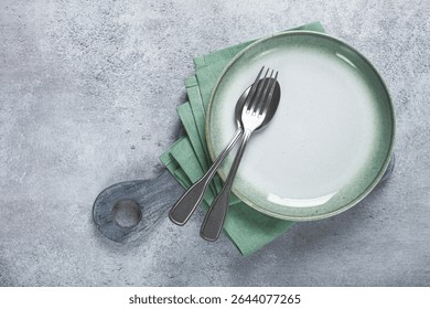 an empty plate and cutlery laid out, on a cutting board, with a green napkin, a gray table, top view,