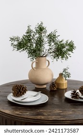 Empty plate with fork and pine cone near ceramic vase jug with green plant branches on wooden vintage table on white background. Serving concept. Decoration element. Prepare for dinner