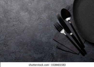 Empty plate, fork, spoon and knife. Black kitchen utensils set on stone table. Top view flat lay with copy space