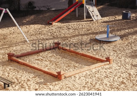 Empty playground in sunlight. Sandpit with brick border. Slide and roundabout nearby. Sunny day at park. No children playing outside. Outdoor recreation area. Public play zone in morning light.