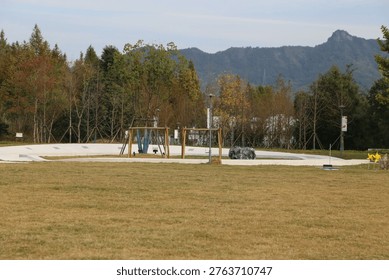 Empty playground with swing set and slide. Trees stand behind the play area. Mountains form a distant backdrop. Grassy field stretches before the equipment. No animals or people are visible