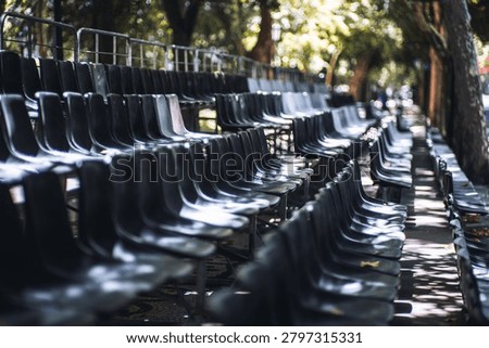 Empty portable city event seating with glossy black plastic chairs under trees, arranged in repeating rows for spectators at an outdoor public venue