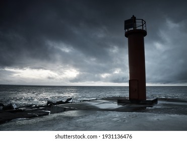 Empty promenade to the red lighthouse with a solar battery, close-up, frozen Baltic sea in the background. Dark storm clouds. Alternative energy, climate change, global warming concepts. Long exposure