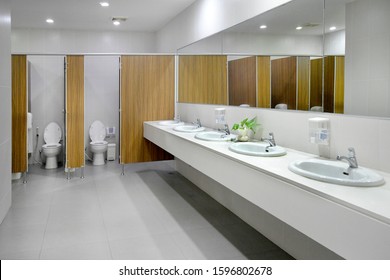 Empty Public Bathroom With White Sinks And Wide Wall Mirror, And Toilets