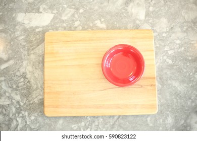 Empty Red Bowl On The Right Side Of A Wooden Cutting Board On Top Of White And Gray Granite Counter Top. Vertical, Overhead Image With Copy Or Text Space.