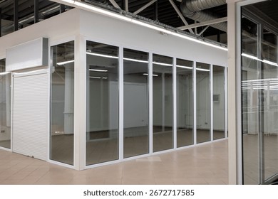 Empty retail unit with closed rolling shutter and glass walls inside shopping mall. Indoor commercial space with tiled floor and blank signboard.