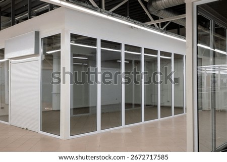 Empty retail unit with closed rolling shutter and glass walls inside shopping mall. Indoor commercial space with tiled floor and blank signboard.