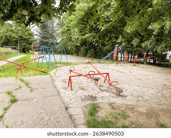 Empty retro playground with seesaws, swings, and a slide under green trees. A quiet and nostalgic public space in the city.