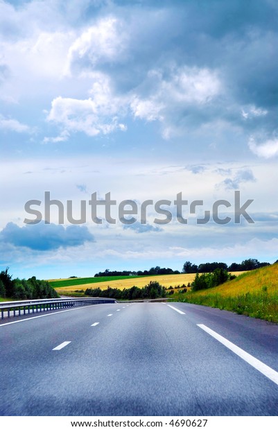 Empty road among\
fields with blue cloudy\
sky