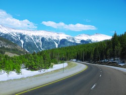 Empty Road In A Beautiful Countryside Of Colorado, USA With Green Pine Trees Along The Road And Mountain Covered With Snow In The Background