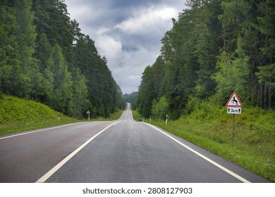 Empty road cutting through dense green forest, framed by tall trees and dramatic overcast clouds. Deer crossing sign warns of wildlife ahead, conveying solitude, travel, safety and moody atmosphere