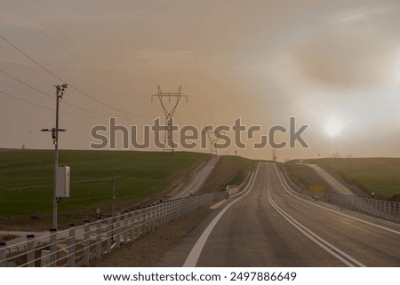 Empty road at sunset in early spring. On the edge of the road a small weather station. A misty, slightly cloudy early spring evening in the hilly suburban areas crossed by an asphalt road. 