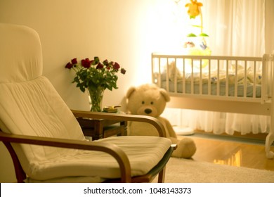 Empty Rocking Chair In A Baby Boy Room With Big Teddy Bear, Crib And Table With Rose Flowers At Night