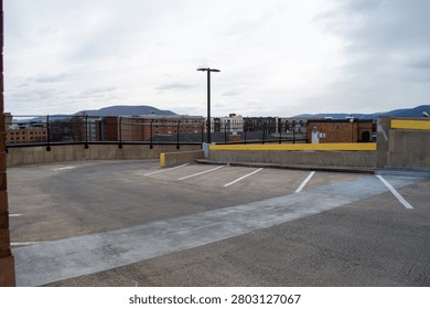 Empty rooftop concrete parking deck featuring white line markings and yellow safety barriers, offering a panoramic view of an urban city skyline with brick buildings and distant mountains under an overcast sky, conveying urban infrastructure and solitude.