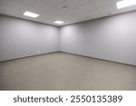 An empty room features plain gray walls and a gray floor. The ceiling has white acoustic tiles with recessed fluorescent lights, creating bright illumination.