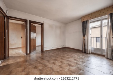 Empty room with old brown stoneware floor, wall air conditioner, cheap wooden access doors to other rooms and balcony with curtains and sheers - Powered by Shutterstock