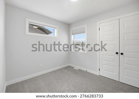 Empty room with soft gray carpet and white walls. Two windows let in natural light. Double doors offer a clean, minimalist entrance. Ceiling light illuminates the entire space