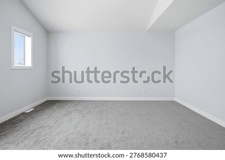 Empty room with soft gray carpet and white walls. Small window lets in natural light, casting gentle shadows. Minimalist space invites imagination or renovation. Ideal for real estate, design