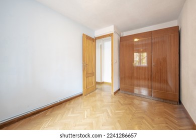 An empty room with a wardrobe with three-section glossy veneered doors with oak parquet floors placed in a herringbone pattern and hot water pipes on the skirting board