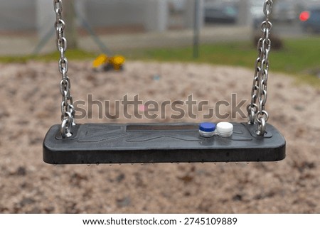 Empty rubber swing with cap resting on seat over sandy playground, shallow depthoffield, muted overcast light, urban park background, evokes nostalgia and quiet solitude, suggests recent
