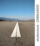 Empty runway ahead with the white line in the midle and a visible crack in it. The grey runway leads to the Pirenees mountains which rests in the background. It is a sunny day with a clear blue sky.