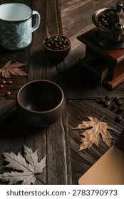 An empty rustic ceramic cup on a dark textured wooden table with a vintage coffee grinder, roasted beans, and dry autumn leaves