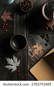 An empty rustic ceramic cup on a dark textured wooden table with a vintage coffee grinder, roasted beans, and dry autumn leaves