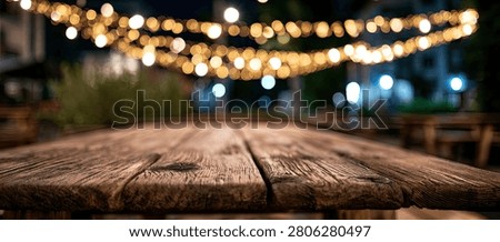 An empty rustic wooden table in the foreground, with blurred bokeh string lights creating a warm, inviting atmosphere in the background at night.